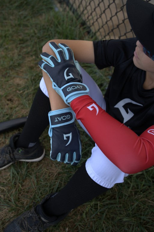 Baseball player sitting near a fence, wearing GOAT batting gloves and a GOAT compression sleeve, resting arms on knees with a focused look.